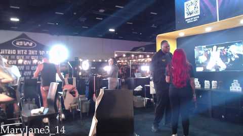 Video of a bustling trade show booth with a red-haired woman in a black dress and a man in a suit, surrounded by colorful displays, bright lighting, and promotional materials.