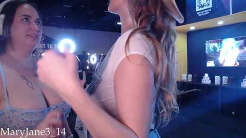 Media: Video of two women in a dimly lit room. One is a light-skinned woman with long wavy hair, wearing a white dress, holding a microphone. The other is a brunette with a tattoo on her chest, wearing a blue dress.