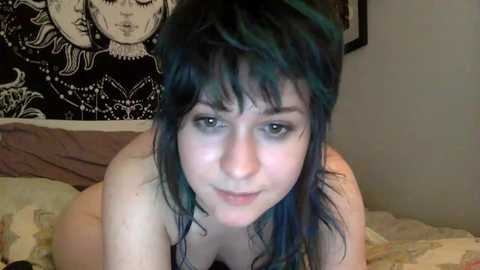 Video of a pale-skinned woman with blue-green hair, lying nude on a bed with a patterned tapestry in the background.