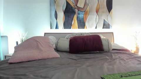 Video of a modern bedroom with a bed dressed in gray and pink bedding, featuring a large abstract art piece on the wall, two white and red pillows, and two bedside lamps.