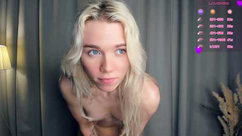 Media: Video of a topless, fair-skinned, platinum blonde woman with blue eyes, leaning forward, in a dimly lit room with grey curtains and a lamp, displaying a chat screen with \"Xxx-Horny\" and \"Xxx-Young\" options.