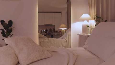 Video of a cozy bedroom with a mirrored closet, white bedding, and a lit lamp on a nightstand; the adjoining kitchen is visible in the mirror.