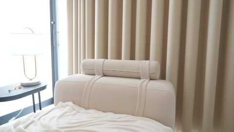 Video of a modern bedroom featuring beige upholstered headboard, matching bedspread, and tall, soft, beige curtains. A white table lamp with a metallic base sits on a round, dark wooden nightstand beside a large window.