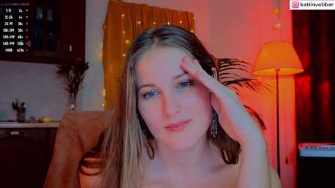 Media: Video of a young woman with long blonde hair, fair skin, and blue eyes, wearing a yellow top, seated in a cozy, dimly lit room with warm, red lighting.