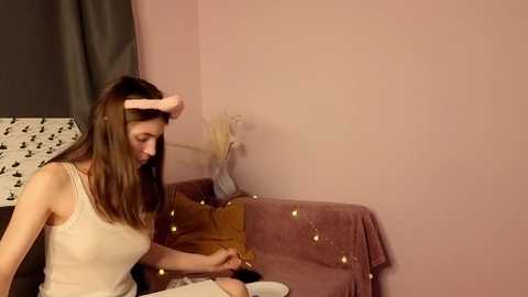 Video of a young woman with straight brown hair, wearing a beige tank top, sitting on a mauve couch with string lights, holding a doll.