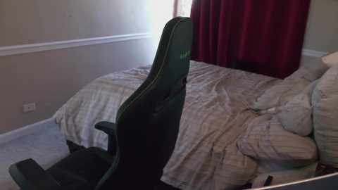 A video of a cluttered bedroom with beige walls, a messy bed covered in light bedding, a black gaming chair, and red curtains partially drawn.