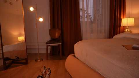 Video of a warmly lit bedroom with a plush bed, beige curtains, and a tall floor lamp. A wooden chair and a mirror are visible.