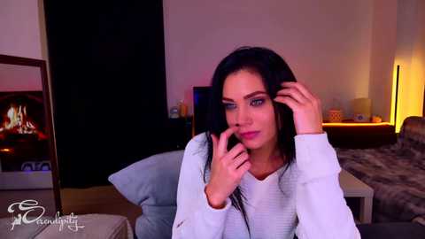 Video of a young, slender woman with long black hair, wearing a light sweater, holding a phone to her ear, seated on a couch in a modern, dimly lit living room with a fireplace and a bed in the background.