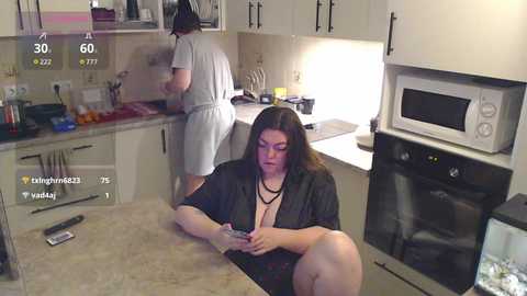 Video of a middle-aged woman with long dark hair, wearing a black robe, sitting on a kitchen counter, using a smartphone. Kitchen background shows another person in a white apron and a microwave.