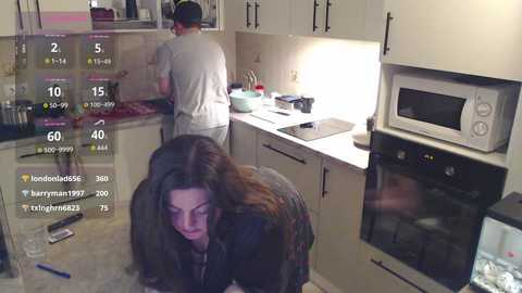 Video of a modern kitchen with beige cabinets and a microwave. A woman with long brown hair kneels on the floor, focused, in a black jacket. A man in a white shirt stands near the sink.