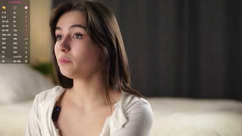 Media: Video of a young woman with long brown hair, fair skin, wearing a white cardigan, looking contemplative in a dimly lit room with a bed and dark curtains in the background.