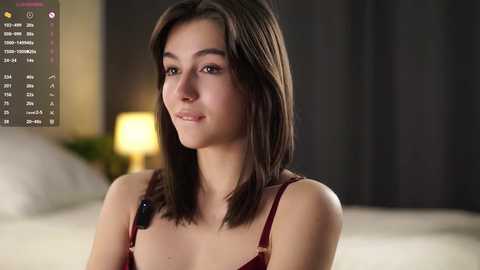 Video of a young woman with straight brown hair, wearing a burgundy camisole, standing in a dimly lit bedroom. Background includes a bed with white linens and a yellow lamp.