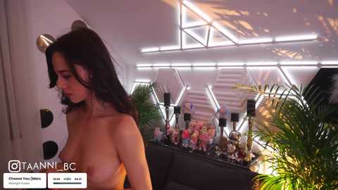 Media: Video of a topless woman with long black hair and medium-sized breasts, standing in a futuristic, modern bar with neon lighting, plants, and bottles.