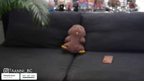 Media: Video of a plush toy resembling a dinosaur on a dark gray sofa, with a blurry background showing a group of people, possibly a crowd.
