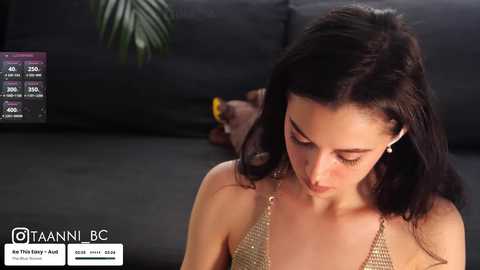 Media: Video of a young woman with long black hair, wearing a gold halter top, sitting on a black couch, with a green leafy plant in the background.