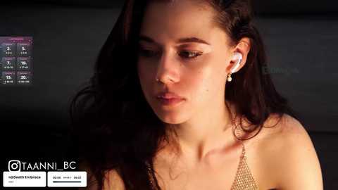 Media: Video of a young woman with fair skin, long dark hair, and a light gold chain necklace, wearing white earbuds, in a dimly lit room with a digital screen in the background.