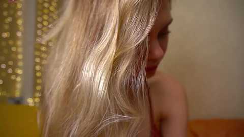 Video of a young woman with long, wavy, platinum blonde hair, partially visible face, and a soft, golden background.