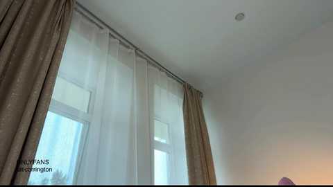 Video of a minimalist room with large, sheer white curtains covering three windows, letting in soft light. The walls are a plain white, with a single small light fixture in the ceiling. The room has a modern, clean aesthetic.