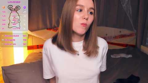 Video of a young, fair-skinned woman with shoulder-length brown hair, wearing a white t-shirt, in a dimly lit room with a bed and a wall-mounted TV displaying \"LOVE ISLAND\" text.