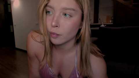 Media: A video of a young, fair-skinned woman with blonde hair, wearing a purple bra, looking contemplative. She has blue eyes and is indoors, in a dimly lit room with a kitchen in the background.