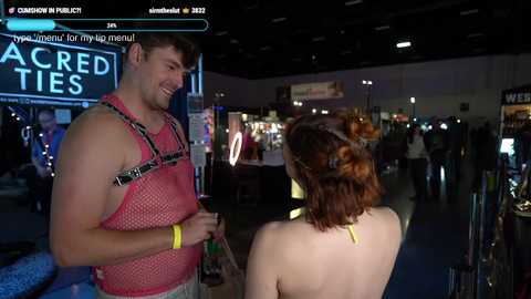 A video of a dimly lit convention hall featuring a man in a red mesh tank top and a woman with auburn hair in a yellow choker, both smiling.