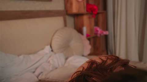 A video of a person with brown hair lying in bed, covered with a white sheet, in a dimly lit room with wooden furniture and a vase of red flowers on a shelf.