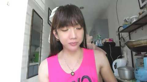 A video of an Asian woman with straight black hair, wearing a pink tank top, making a pouty face in a cluttered room with shelves, containers, and a mirror.