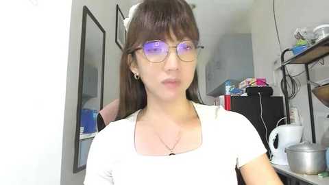 Video of an Asian woman with long brown hair, wearing glasses, a white top, and a necklace, standing in a cluttered kitchen with white walls, shelves, and appliances.