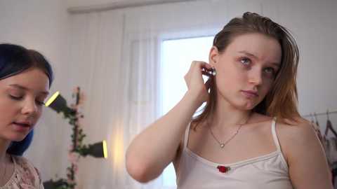 Video of a young woman with light skin and long brown hair, wearing a white tank top, adjusting her earrings in a well-lit, modern room with white walls and curtains.