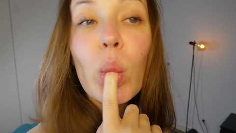 Video of a Caucasian woman with straight, shoulder-length brown hair, light skin, and blue eyes, playfully placing her index finger on her lips. Background includes a white wall, a lamp, and a hint of furniture.