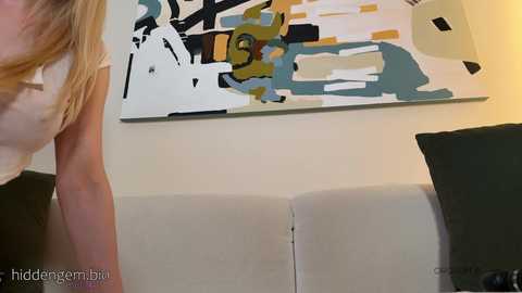 Media: Video of a blonde woman in a white top, standing in front of a modern abstract painting with bold, geometric shapes in black, white, and yellow on a beige wall, and a gray couch.