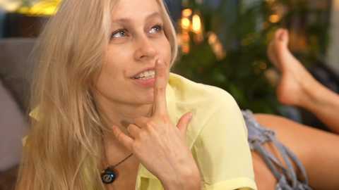 Media: Video of a smiling, fair-skinned, blonde woman with long hair, wearing a yellow blouse, lying on her stomach on a couch.