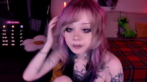 Video of a young woman with pale skin, purple hair, and numerous tattoos, including a spider web on her chest, in a dimly lit bedroom with a calendar, plush toys, and a plaid bedspread.