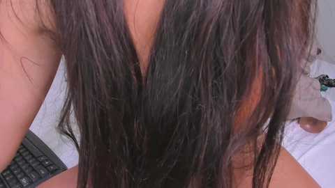 A close-up video of a person with long, straight black hair parted down the middle, revealing a smooth, light-toned neck. The background shows a white towel and a blurred object.