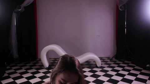 A video of a young woman with long brown hair lying on a black and white checkered floor, surrounded by white, abstract, curving objects in a dimly lit, surreal room with red and black walls.