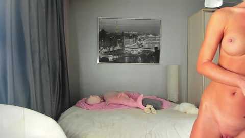 Video of a naked man in a modern bedroom with a gray wall, a black-and-white framed photo, a white lamp, and a messy white bed with pink sheets and toys.