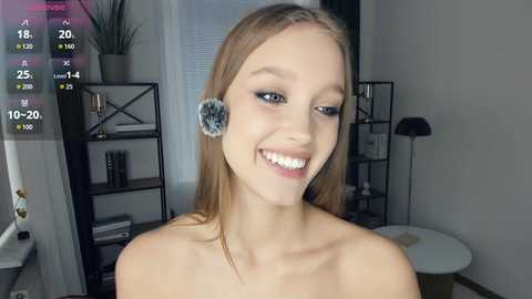 Video of a smiling young woman with straight, light brown hair, wearing fluffy blue headband earrings, in a modern, minimalist room with shelves and a black floor lamp.