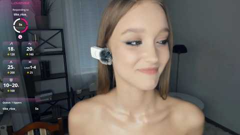 A video of a young, light-skinned, blonde woman with straight hair, wearing a white and black headband, smiling in a modern, minimalistic room with a grey wall and a bookshelf.