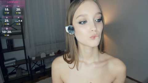 Video of a young, fair-skinned woman with straight, light brown hair and light makeup, wearing a fluffy, round hair accessory. She's topless, indoors, with a background featuring a black bookshelf and white curtains.