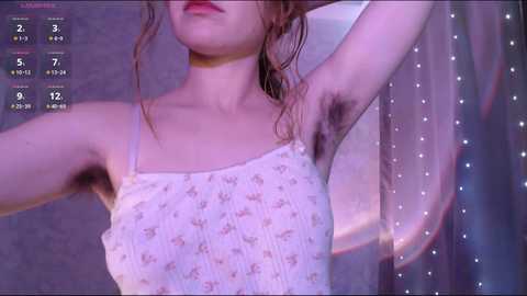 Video of a young woman with light skin, wearing a white floral-patterned tank top and a choker. She has wet, tousled hair and armpit hair visible. Background includes a mirror and a curtain.