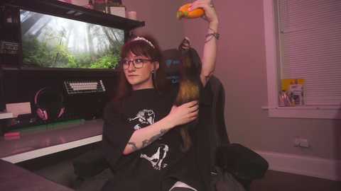 Video of a young woman with glasses and dark hair, wearing a black T-shirt and holding a large snake, seated in a gaming chair in a room with a TV, gaming equipment, and a window.