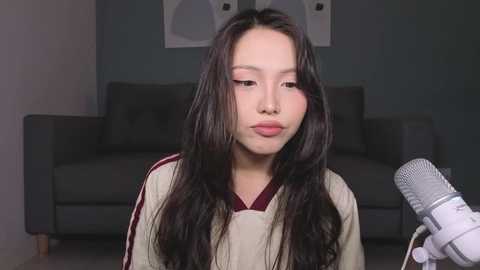 Video of a young Asian woman with long, wavy black hair, wearing a beige sweater with red trim, sitting in front of a grey couch, holding a grey microphone.