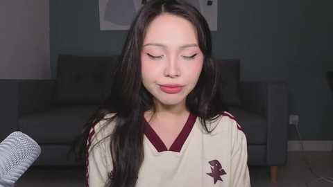 Video of a young Asian woman with long black hair, wearing a white shirt with burgundy accents, sitting in a modern living room with a grey couch and light grey walls.