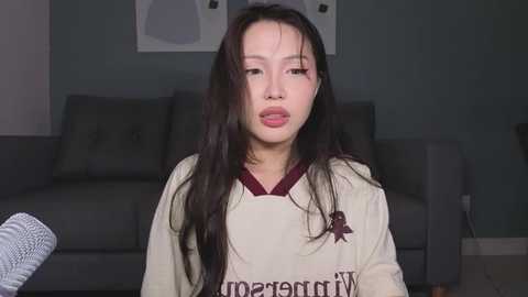 Video of an Asian woman with long black hair, wearing a white sweater with a red logo, sitting in a living room with a gray sofa and abstract art on the wall.