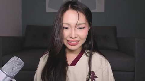 A video of an East Asian woman with long black hair, wearing a beige kimono, smiling, with a microphone in the foreground, and a dark gray couch in the background.