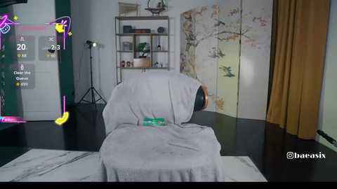 Video of a cozy, modern living room with a plush, grey armchair draped in a white blanket. A tall, green plant sits on a wooden shelf, and beige curtains frame the background.