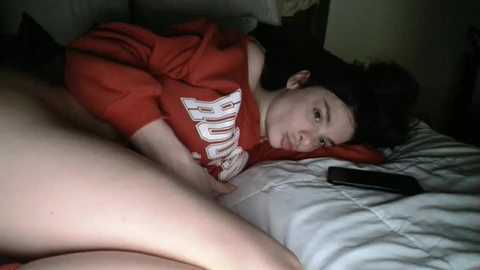 Media: Video of a young woman with fair skin, lying on her side on a bed, wearing a red \"Baylor\" T-shirt, black hair, and a smartphone beside her.