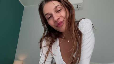 Video of a young woman with long, wet, brown hair, closed eyes, and slight smile, wearing a white, long-sleeved top, in a modern, minimalist room with green and white walls.