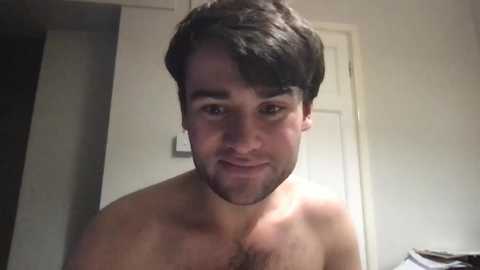 A video of a shirtless, bearded Caucasian man with short dark hair, wearing a faint smile, standing indoors against a plain white wall and door background.