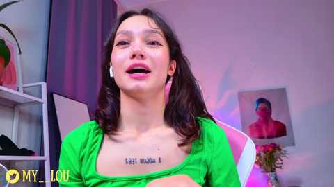 Video of a young woman with medium skin tone, shoulder-length dark hair, wearing a green top with a tattoo reading \"I am not a boy\" on her chest, smiling. Background includes a purple curtain, white shelves, and a pink flower vase.